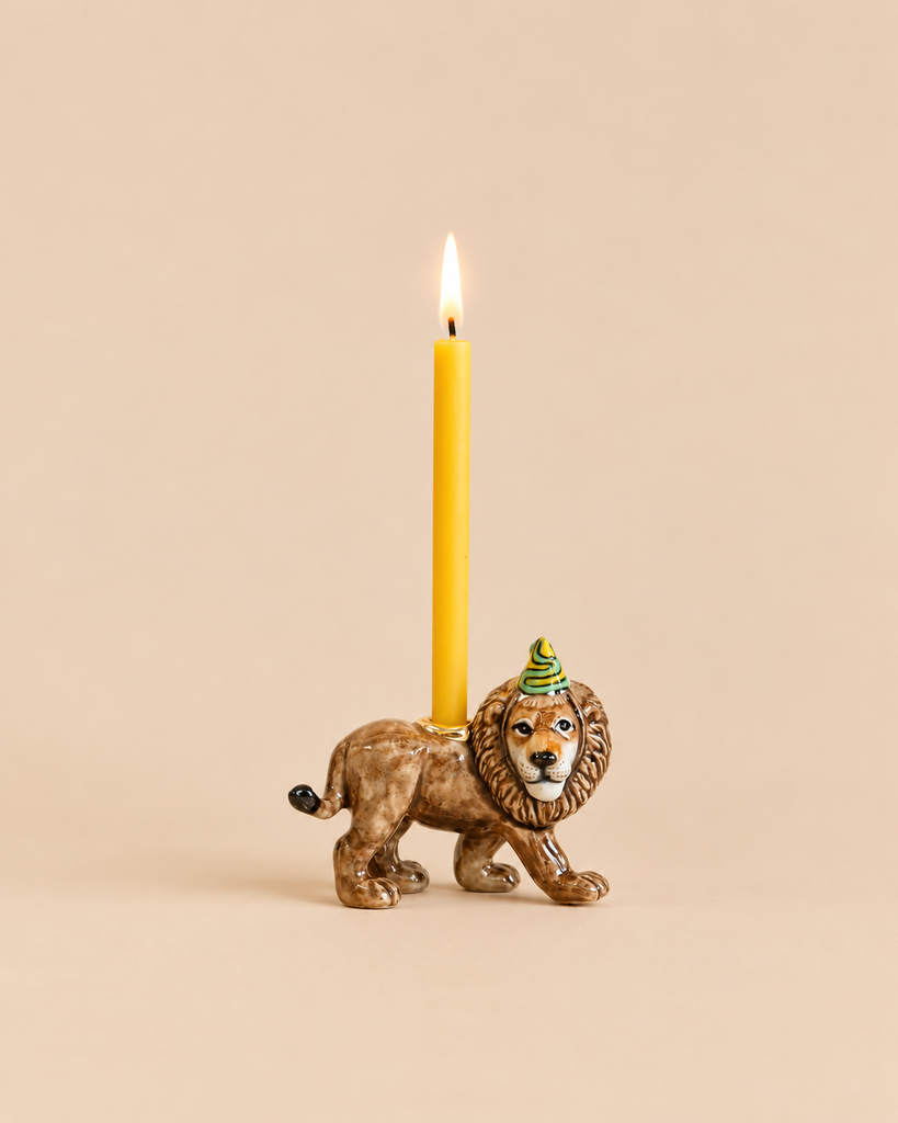 A Lion Cake Topper - Final Sale handcrafted as a ceramic lion figurine wearing a party hat, featuring a holder on its back with a lit yellow taper candle.