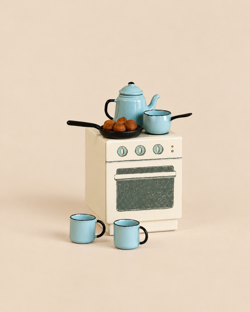 A Maileg Madam Blue’s Favorites toy kitchen stove topped with a blue teapot, a small saucepan, and a frying pan containing small brown treats, with two blue mugs on the floor in front.