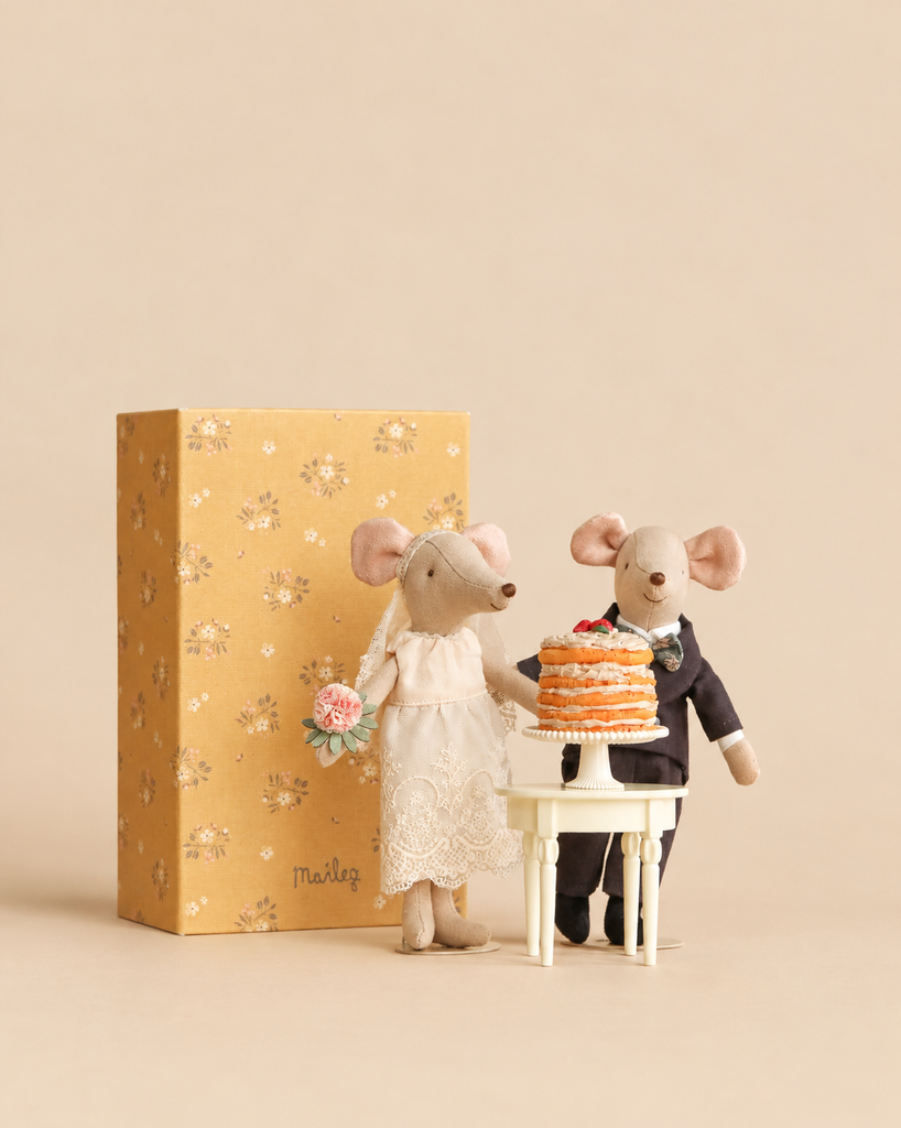 Maileg Wedding Day Set mice, dressed as a bride and groom, stand beside a party cake on a pedestal table next to a patterned gift box.