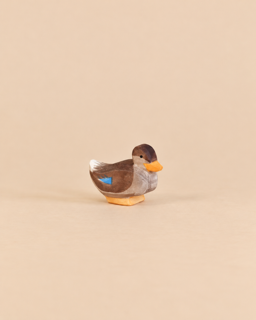 A small handcrafted wooden Ostheimer Duck - Sitting in a sitting position with brown feathers, an orange beak, and a blue patch on its wing.