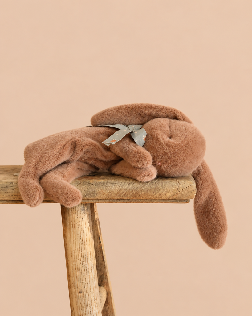 A soft brown Maileg Plush Sleeping Bunny with a small ribbon around its neck resting on a wooden stool.