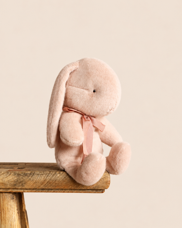 A small, light powder Maileg Plush Bunny with a matching satin ribbon around its neck, sitting on the edge of a wooden stool.