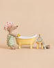 A stuffed mouse wearing a shower cap and towel stands by a yellow Maileg Bathtub Set with a rubber duck, a stool, a small bottle of shampoo, and a hairbrush.