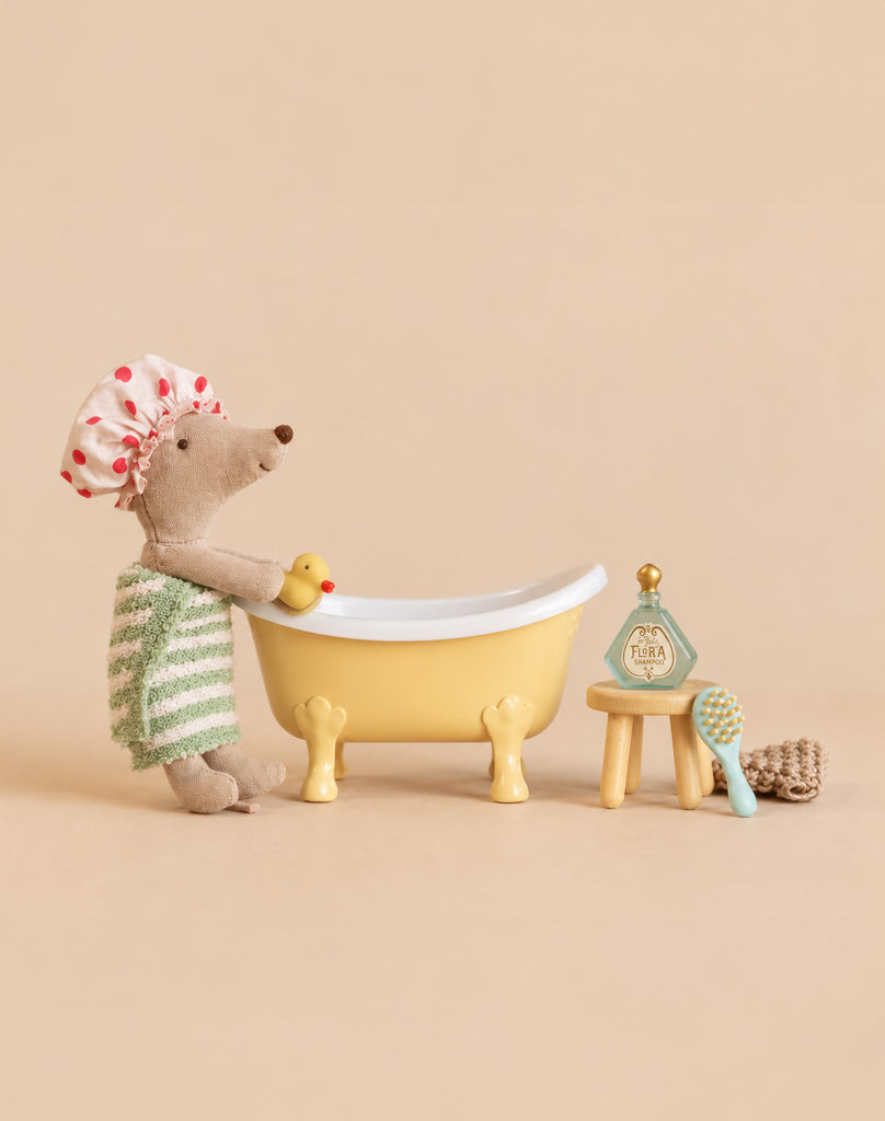 A stuffed mouse wearing a shower cap and towel stands by a yellow Maileg Bathtub Set with a rubber duck, a stool, a small bottle of shampoo, and a hairbrush.