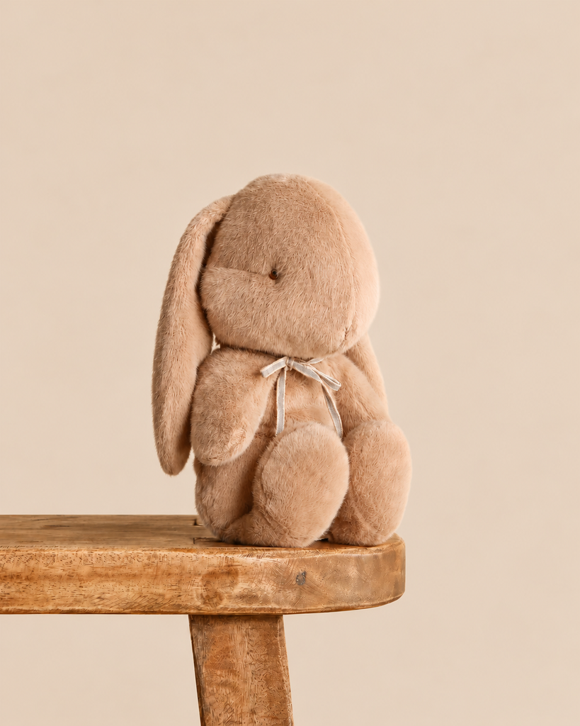 Maileg Plush Bunny, Medium - Cream Peach stuffed animal with long ears and a thin ribbon tied around its neck, sitting on a wooden stool.