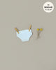 A pair of white cotton Minikane Doll Clothing | Culotte in White Cotton is clipped to a string with two wooden clothespins. To the right, there are two small yellow flowers with thin stems. In the upper right corner, a gray circle reads, "fits MINIKANE 11,13,14"." The background is a light beige color.