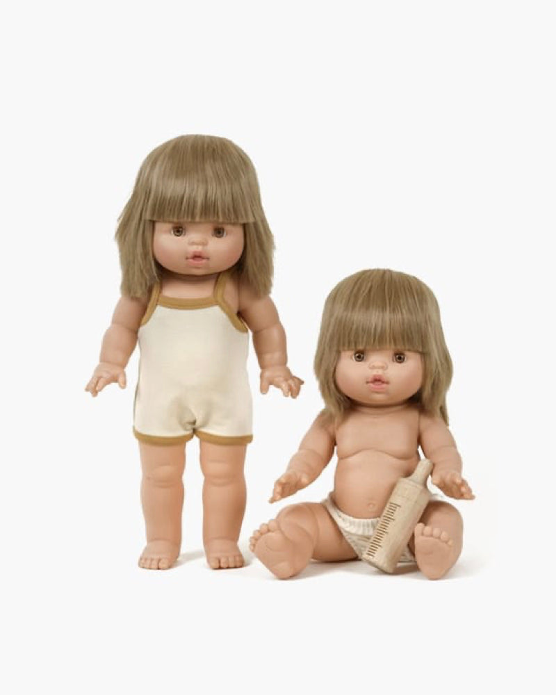 Two Minikane baby dolls with straight, light brown hair and bangs. The 14" standing Zoelie doll wears a cream sleeveless outfit, while the seated doll in a cream diaper holds a wooden comb. Both are anatomically correct, share similar facial features, and have a hint of vanilla scent.