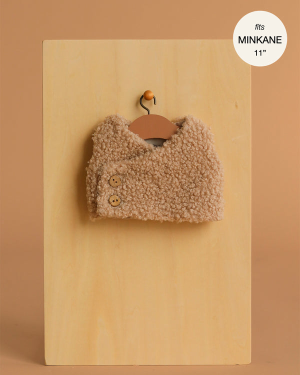 The Minikane Doll Clothing | Babies – Terry Sleeveless Vest in beige is a small, sleeveless vest with two buttons, displayed on a hanger against a tan background. Fits Minikane 11'' dolls—perfect for baby doll outfits.
