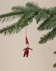 The Maileg Pixy Ornament - Boy, a small fabric elf in red clothes, hangs from a string on the tip of a green pine branch against a plain beige background.
