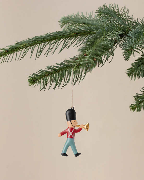 A Christmas tree branch displays the Maileg Metal Ornament, Guard - Trumpet: a hand-painted toy soldier in a tall black hat, red coat, and blue pants playing a gold trumpet. The background is plain and light-colored.