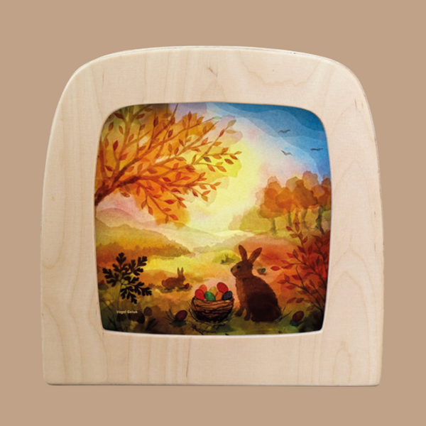 The "Silhouette Only - Easter Basket for Toverlux Lamp" features a vibrant illustration in a wooden frame, showcasing an autumn landscape with foliage, a rabbit on the grass, and a basket of eggs. Crafted from translucent material, its warm hues create Magic Silhouettes that enhance its tranquil charm.
