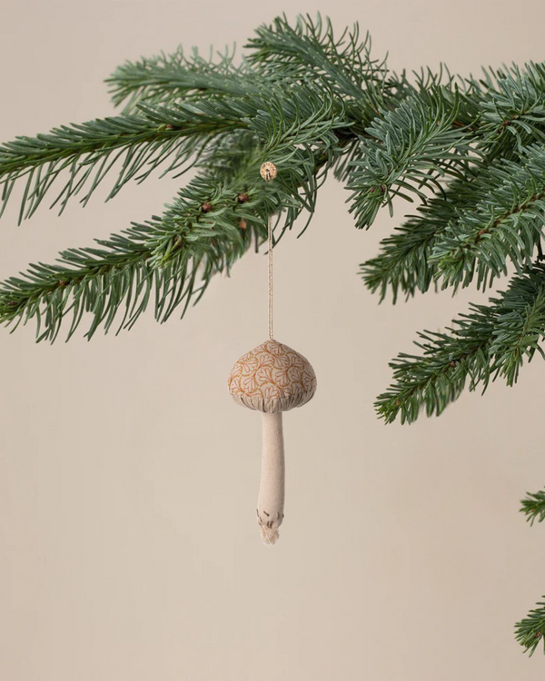 A close-up of a green pine branch with the Maileg Christmas Fabric Ornament, Mushroom - Winter Heart, hanging from it, highlighting the charm of unique holiday decor against a simple, light background.