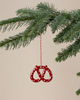 The Maileg Christmas Metal Ornament - Pretzel - Dot, a red hand-painted pretzel with white dots and a red-and-white hanging string, is displayed on a green pine branch against a plain beige background.
