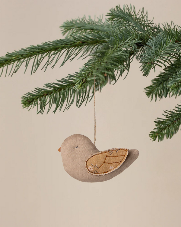 The Maileg Fabric Ornament, Bird - Mable - Latte features stitched wings and hangs from a string on a pine branch, making it a charming addition to Christmas tree ornaments against a neutral background.