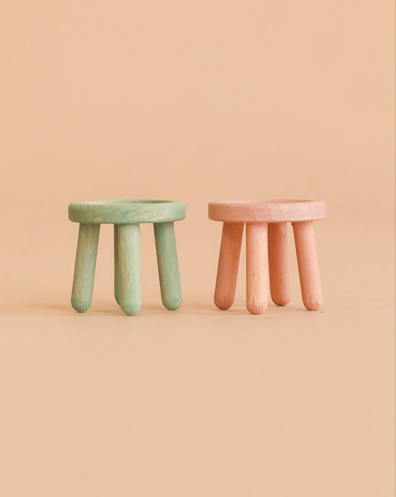 Two small, round, three-legged stools—one green and one pink—from the Maileg Suitcasa Furnished Set are placed side by side on a plain beige background, making them charming additions to any Maileg mouse furniture collection or delightful Maileg dollhouse accessories.
