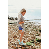 A young girl smiles on a rocky beach, hugging the Senger Naturwelt Cuddly Animal - Whale handmade in Germany. She wears blue sandals, blue shorts, and a striped shirt, with the ocean and cloudy sky in the background.