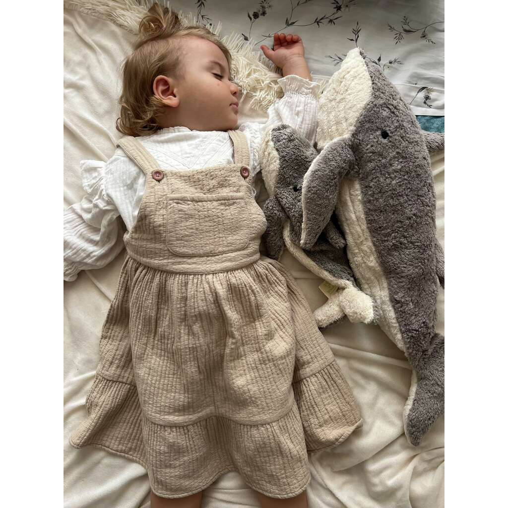 A young child with light hair sleeps on a bed, wearing a beige dress and white top, holding the Senger Naturwelt Cuddly Animal - Whale. She rests on light-patterned organic cotton bedding for extra comfort.