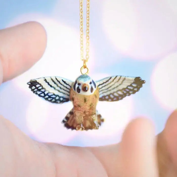 The limited-edition Kestrel Falcon Necklace features a handcrafted porcelain bird pendant with outstretched wings, suspended from a gold chain and showcased between two open hands against a softly blurred background.