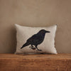 The Coral & Tusk Blackbird with Acorn Pillow is Oeko-Tex certified linen, beige in color, with an embroidered blackbird holding a branch, displayed on rustic wood against a tan wall.