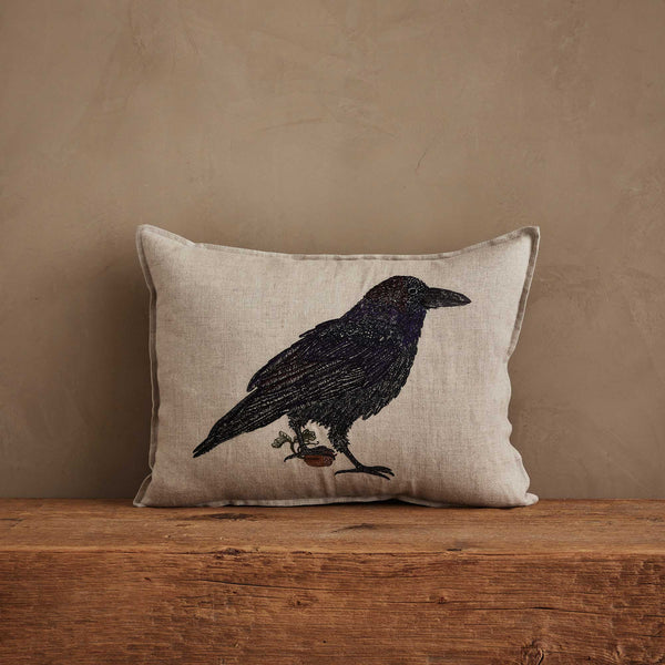 The Coral & Tusk Blackbird with Acorn Pillow is Oeko-Tex certified linen, beige in color, with an embroidered blackbird holding a branch, displayed on rustic wood against a tan wall.