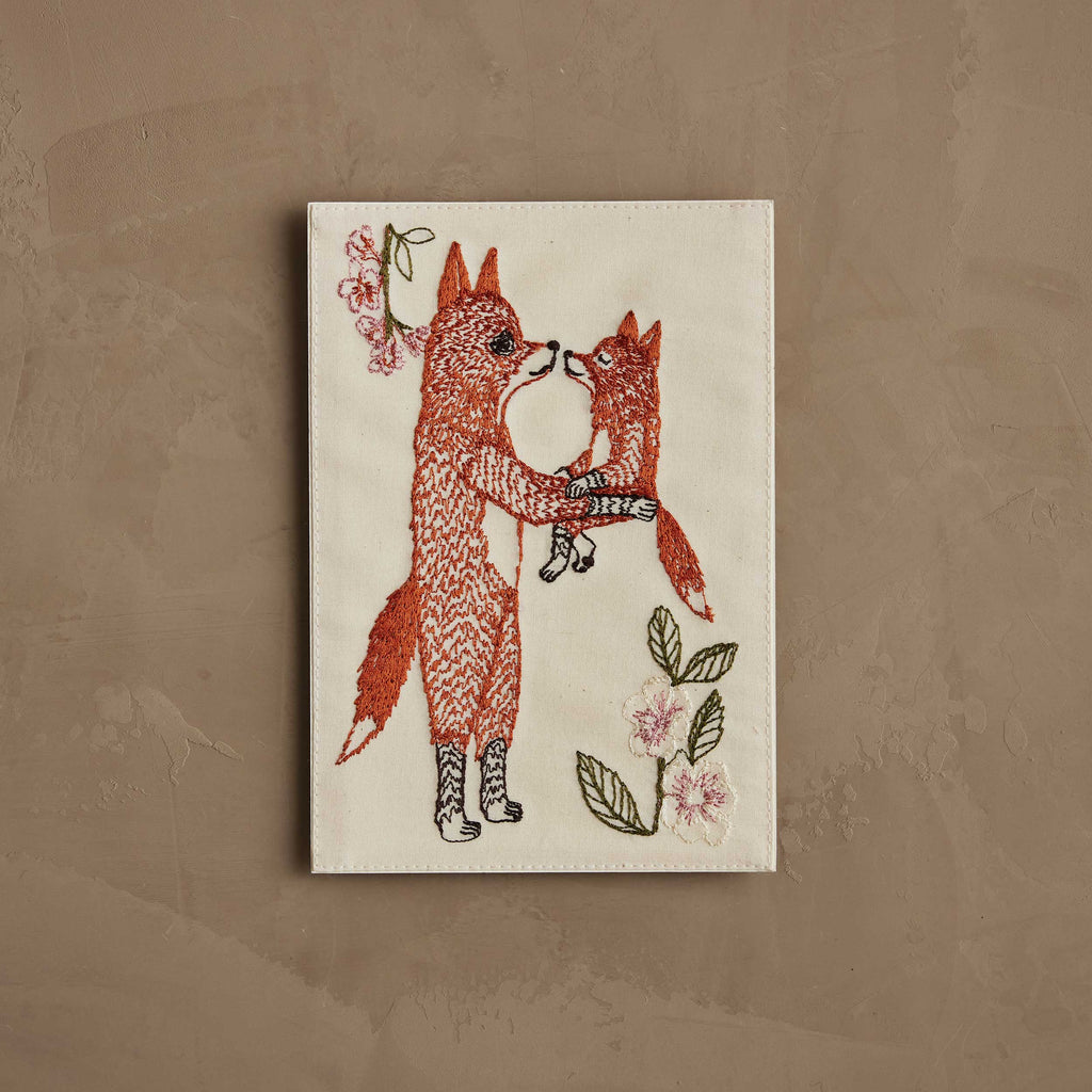 The Coral & Tusk Fox Mama Card features embroidered art of an adult fox hugging a smaller fox, surrounded by pink flowers and green leaves on beige fabric—perfect as a heartfelt new parent gift or charming keepsake.