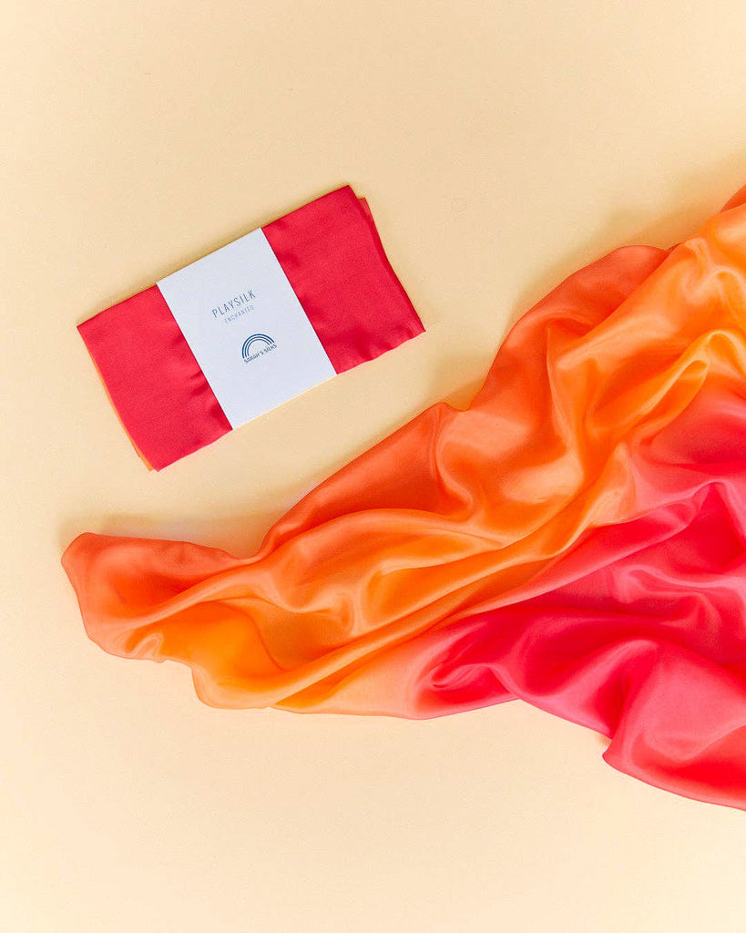 Sarah's Silks Enchanted Playsilk - Fire, made of pure silk in gradient orange and pink, is spread out on a beige surface for open-ended play, with a neatly folded section and white label beside it.
