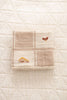 The Organic Cotton Baby Blanket - Farm, in beige and cream patchwork with embroidered chickens and a yellow house, is folded on a quilted off-white surface.