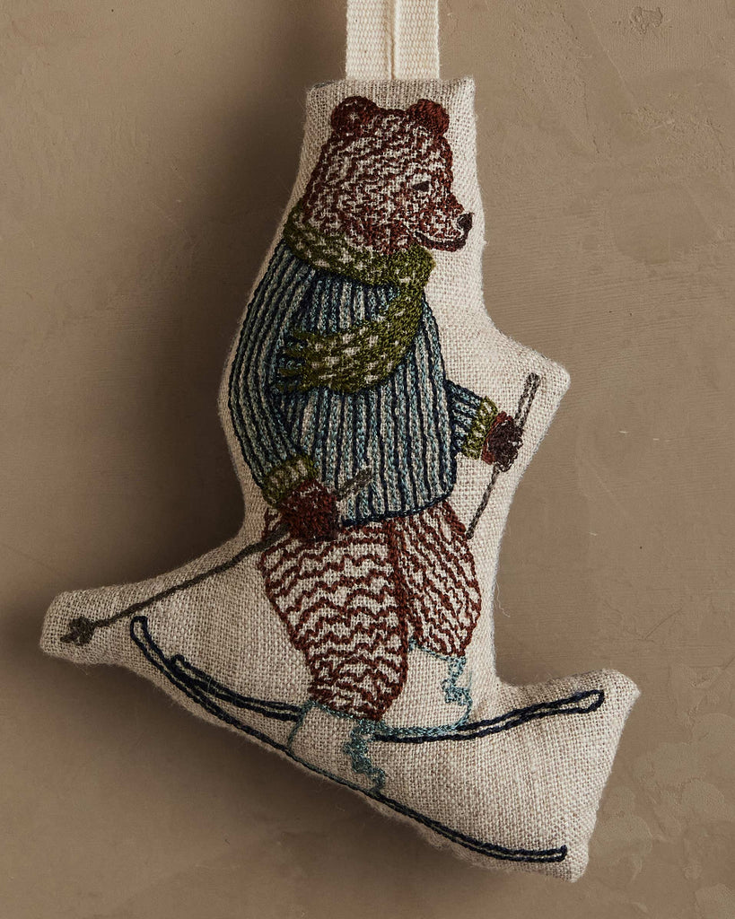 The Coral & Tusk Downhill Bear Ornament features a bear skiing with poles, embroidered in a green scarf, blue sweater, and gloves. It hangs from a loop, making it an ideal holiday or winter decoration for any backdrop.