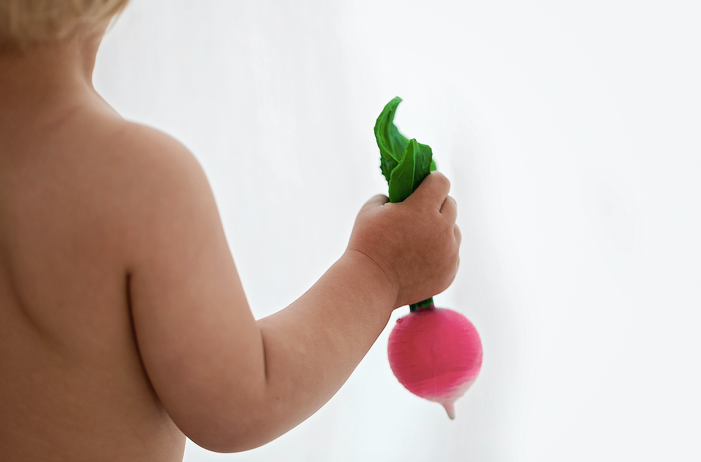 A young child with bare shoulders holds Ramona the Radish Teething Toy—a small pink teether with green leaves—against a plain, light background, perfect for sensory play.