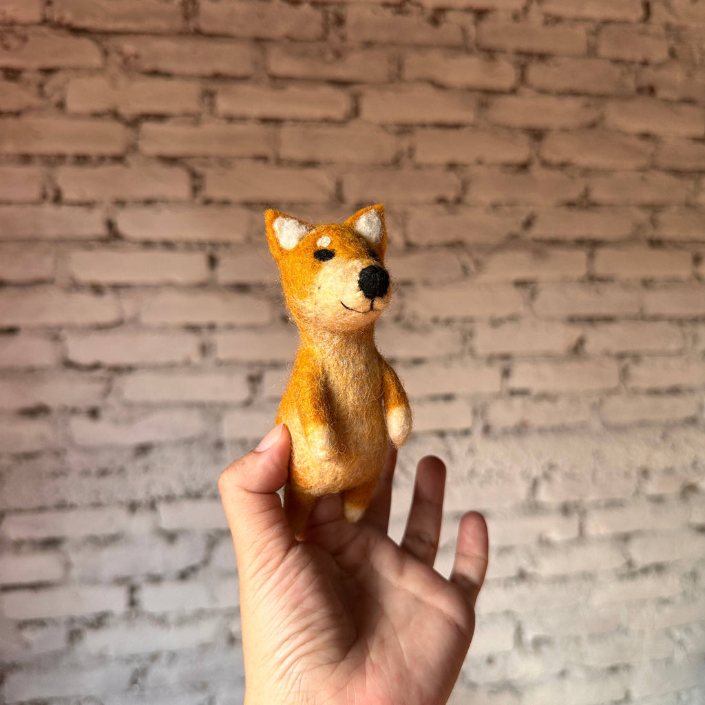 A hand holds up a small, handmade Shiba-Inu Dog Finger Puppet crafted from wool against a white brick wall background. The orange puppet features charming white accents and a friendly expression.