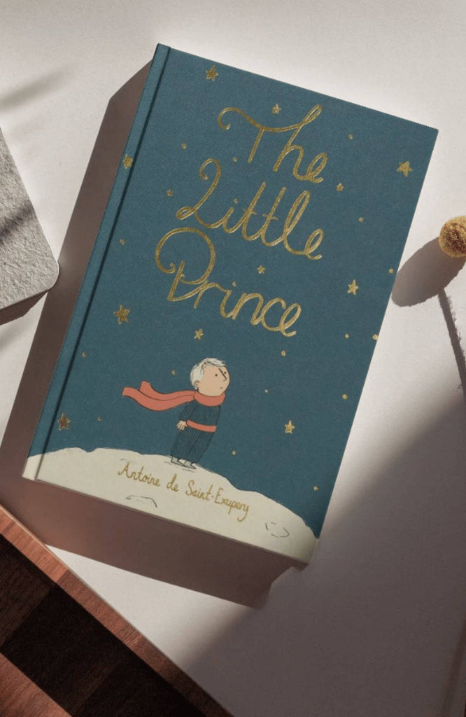 A copy of The Little Prince | Collector’s Edition | Hardcover rests on a white surface, showing the prince with a red scarf beneath gold stars on deep blue—a modern fable by Antoine de Saint-Exupéry.