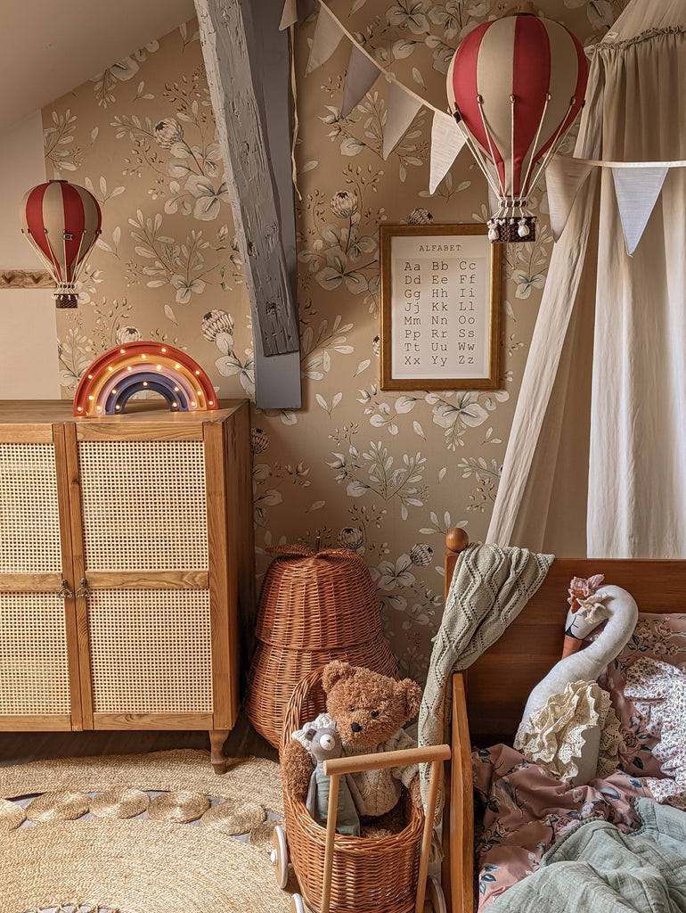 A cozy children’s bedroom with floral wallpaper, the Hot Air Balloon - Burgundy wooden decor, a rattan wardrobe, a wicker basket, rainbow light, bunting, and a wooden bed with plush toys and cushions—perfect for charming children's room decoration.