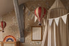 A cozy children's room features floral wallpaper, a burgundy hot air balloon decor, a framed alphabet chart, a fabric canopy, white pennant banner, and a colorful rainbow nightlight on a rattan cabinet.