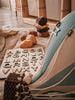 A wooden alphabet puzzle board with letters A-Z sits on a beige surface beside stacked blocks, a blue-striped padded chair, and a mint green and grey Hot Air Balloon decoration in a cozy, sunlit room.