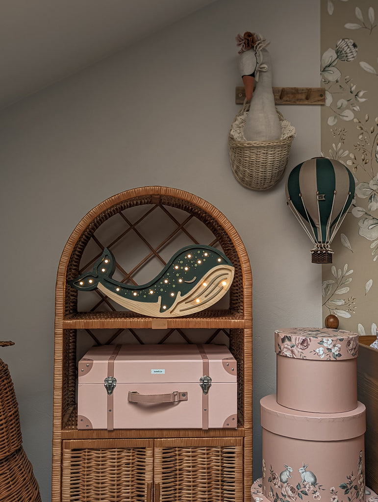 A cozy nursery corner features a rattan shelf with a light-up whale, pink suitcase, and floral hat boxes. Above it hang a fabric horse head in a basket and the Hot Air Balloon - Light Brown / Green children's room decoration.