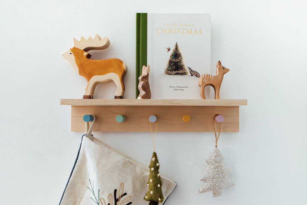 A wooden shelf displays "Our Little Adventure at Christmas (Baby's first Christmas)," wooden animal figures, and festive tree and snowflake ornaments—perfect holiday gifts to inspire toddler language development against a white wall.
