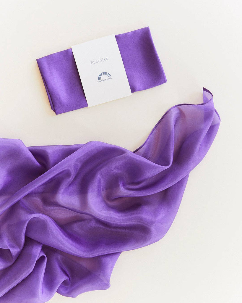 A folded and packaged Sarah's Silks Solid Color Playsilk - Purple sits atop a larger, flowing purple section of the same fabric on a light background. The label reads "PLAYSILK" with a rainbow—perfect for open-ended play.