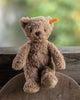 The Steiff Thommy Teddy Bear Brown Plush Toy, 8 inches, with Steiff’s signature yellow tag in its ear, sits on a wooden surface in front of a rustic fence and a softly blurred green and yellow background.