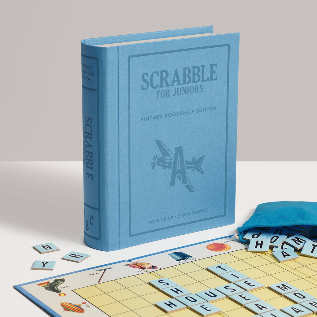 A blue Scrabble for Juniors Vintage Bookshelf Edition game box standing upright next to a partially played children’s word game board, letter tiles, and a blue drawstring pouch.
