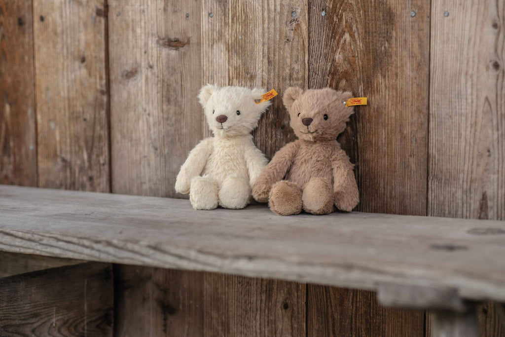 A Steiff Thommy Teddy Bear Brown Plush Toy, 8 inches, sits on a rustic wooden bench. The bear has a signature orange ear tag—a hallmark of Steiff’s quality and tradition.