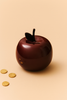 The Ferm Living Apple Money Bank, in glossy dark red with a coin slot on top, sits on a light beige surface beside three gold coins—a stylish and charming way to save your change.