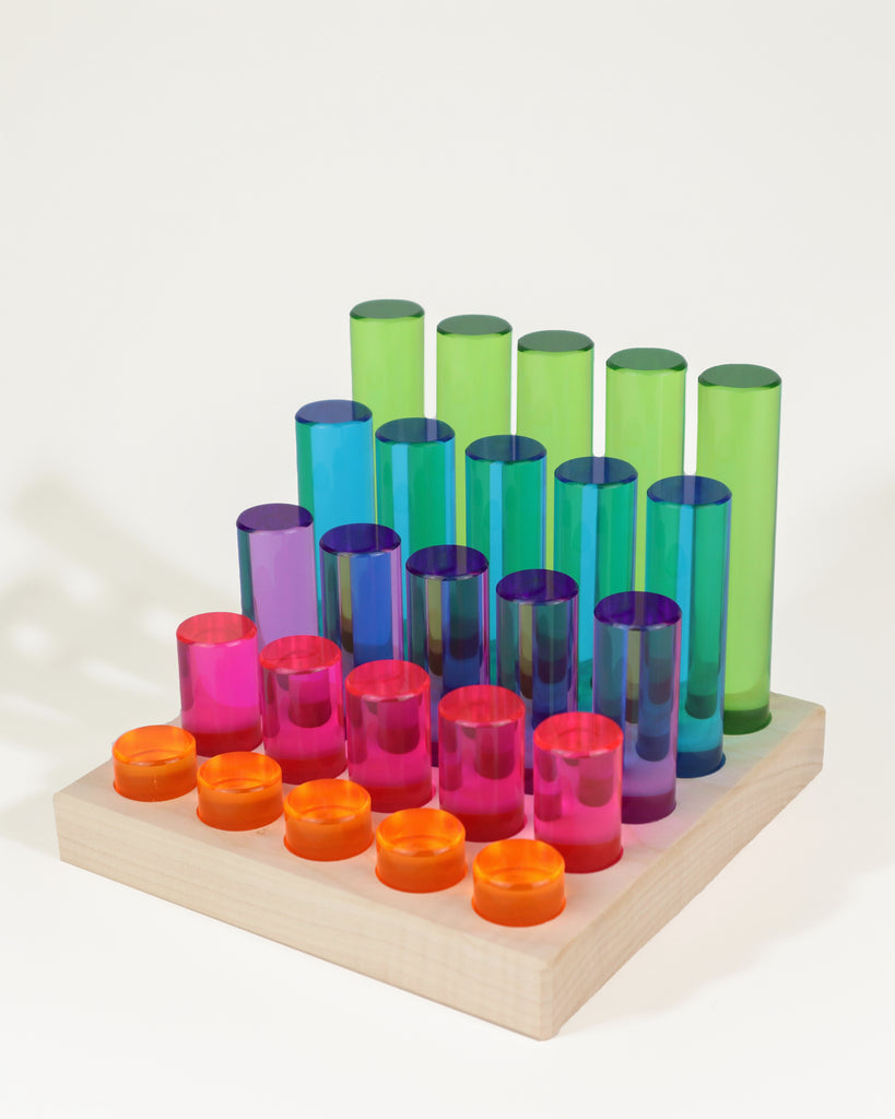 Bauspiel Luminous Pillars 25-Piece Set consisting of twenty-five translucent, colored cylindrical pillars of varying heights arranged in rows on a wooden base.