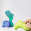A hand holds a small, green, handpainted wooden piece from Grimm's Casa Aqua Building Blocks. In the blurred background, more blue and teal blocks are stacked against a white surface, inviting imaginative play.