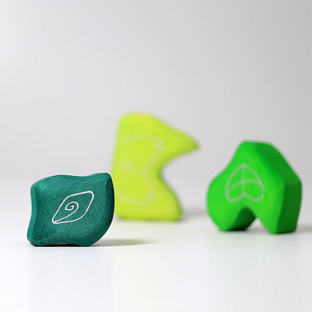 Three abstract, green and yellow Grimm’s Tower Leaves Building Blocks with white line designs are arranged on a white surface, featuring one dark green block in focus in the foreground and two lighter blocks blurred in the background.