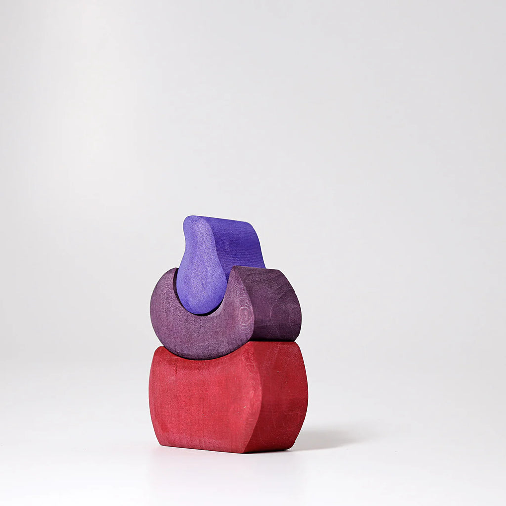 Grimm's Small Bakery Building Blocks: minimalist sculpture of three stacked, smooth wooden blocks in red and purple tones, set against a plain white background.
