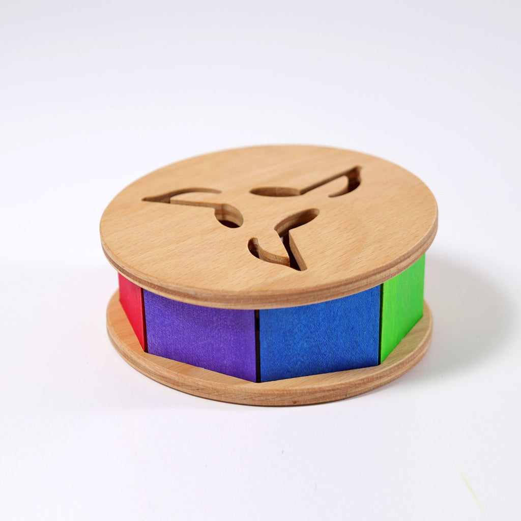Grimm's Sound and Color Wheel is a round wooden baby toy featuring colorful red, blue, purple, and green panels with cut-out shapes on top, shown against a white background.