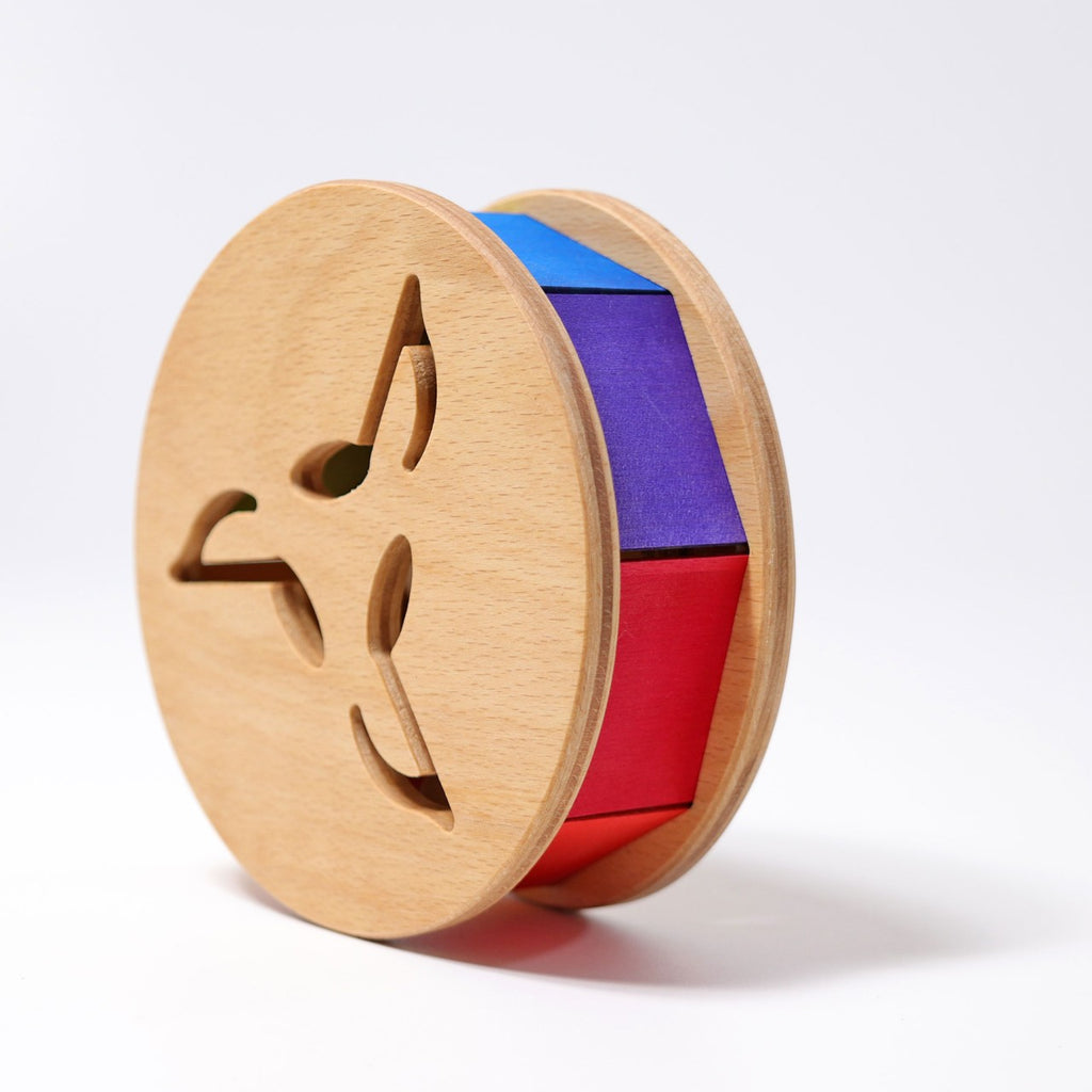 Grimm's Sound and Color Wheel is a non-toxic wooden toy featuring triangular cutouts on each side with red, purple, and blue fabric panels in the center—an engaging motor skill toy for babies shown on a plain white background.