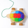 A Grimm's Rainbow Turtle Pull-Toy and Building Blocks with colorful removable geometric pieces on its shell, designed to develop fine motor skills. The turtle has four wheels and a green string for pulling along. Crafted from non-toxic materials, the pieces come in shades of blue, green, red, and orange, creating a rainbow effect.