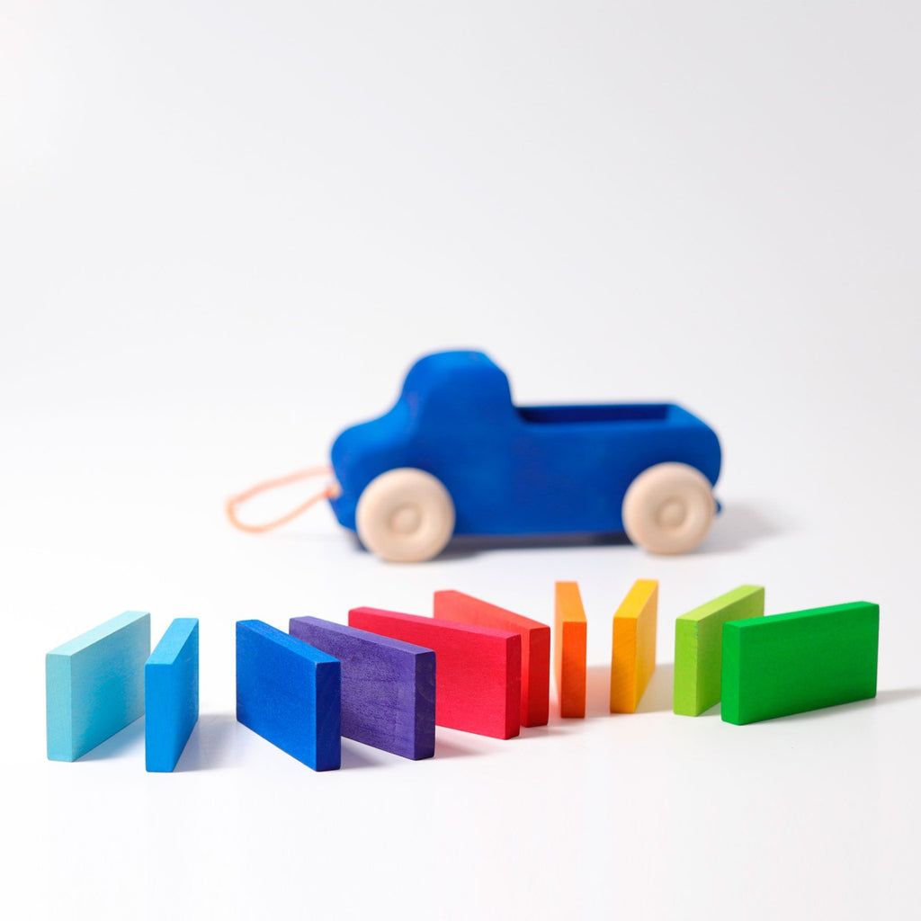 The Grimm's Pull Along Truck, a rainbow-colored toy car with white wheels and a string, is set against a plain white background behind vibrant wooden blocks resembling dominoes about to topple from blue to green, all crafted with non-toxic materials.