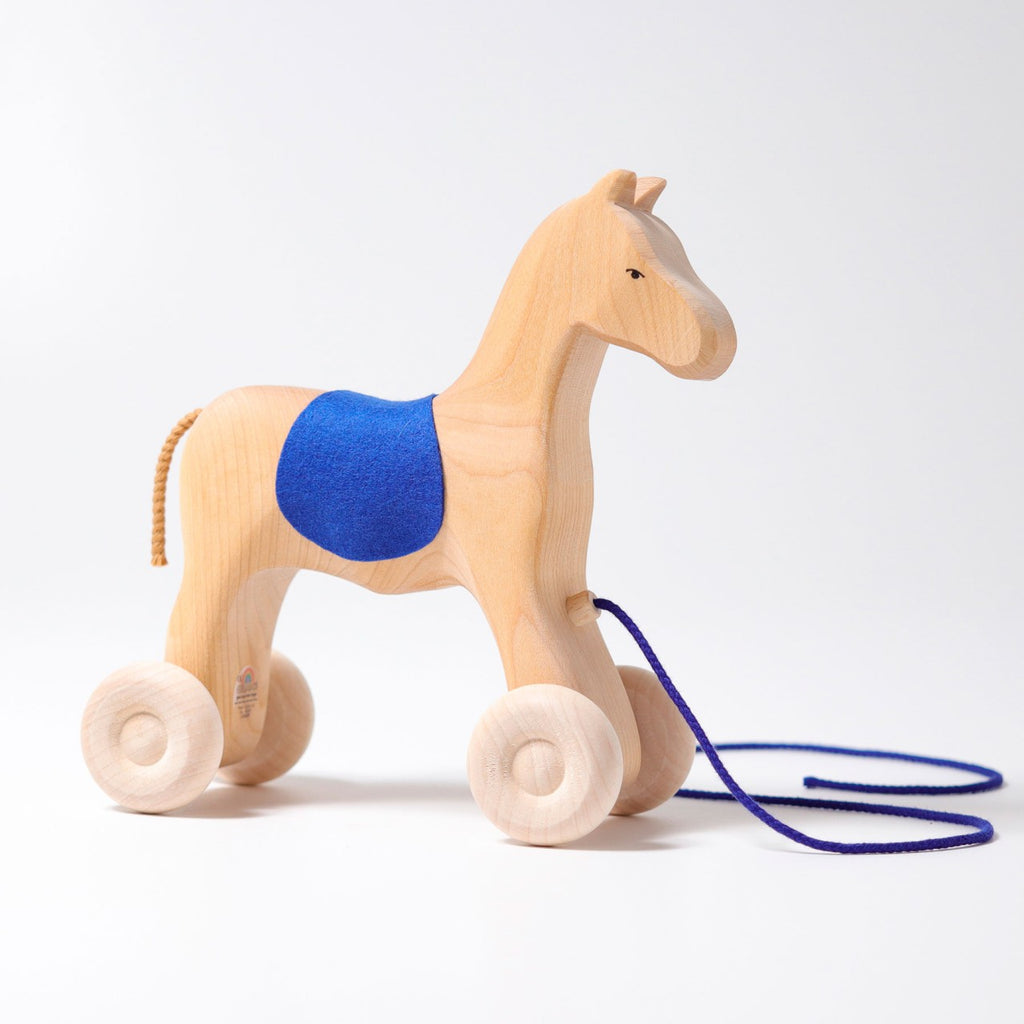 Grimm's Horse Holly is a charming wooden pull-along horse featuring a blue saddle, four wheels, and a blue pull string—perfect as a toddler toy. Set against a plain white background.